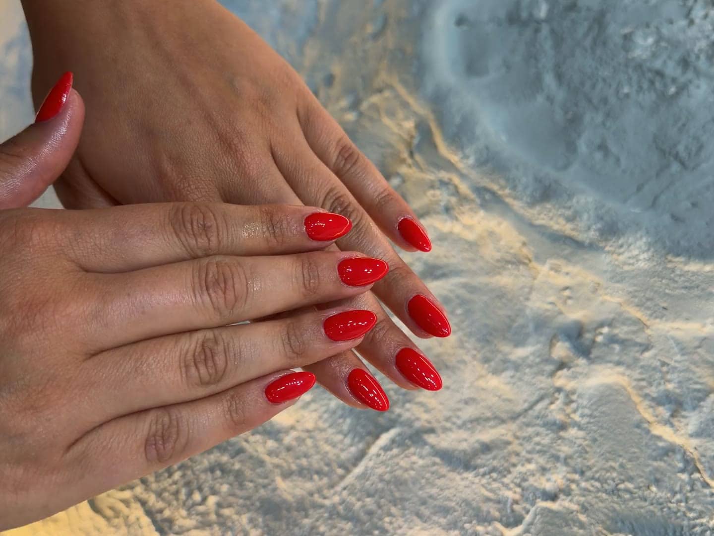 Glossy red almond nails against a textured wall in warm evening light, done at Em Nail Studio in Oakville
